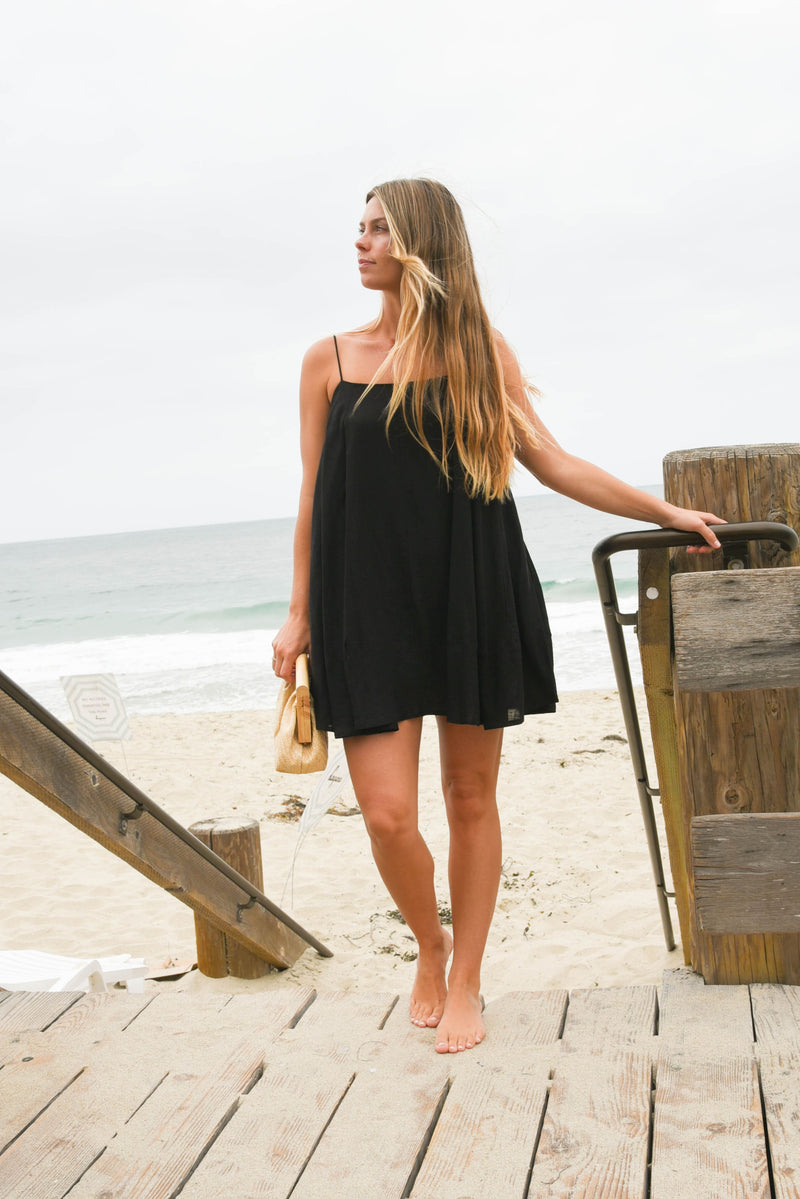 Woman in a black dress standing on a wooden boardwalk by the beach.